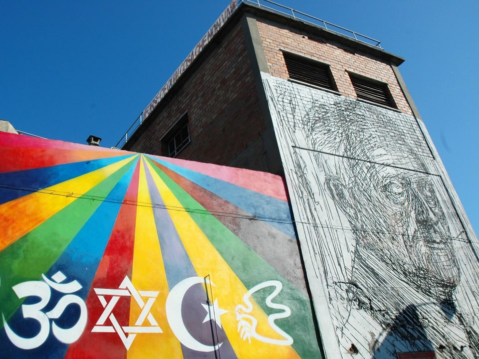 Eine mit Symbolen der Weltreligionen, einem Porträt und bunten Strahlen bemalte Fassade.