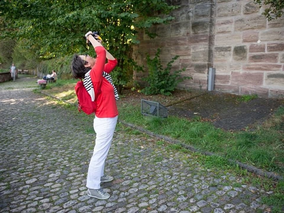 Eine Frau mit weissen Hosen und rotem Pullover fotografiert eine Mauer. 