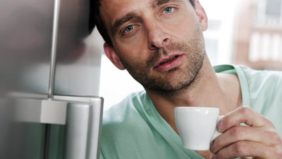 Mann mit müdem Gesicht und einer Espressotasse in der Hand lehnt an einen Kühlschrank