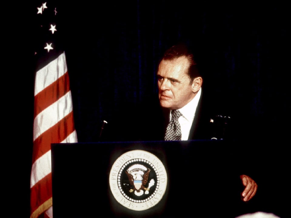 Der Schauspieler Anthony Hopkins steht als Nixon neben einer amerikanischen Flagge am Rednerpult.