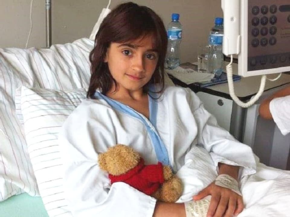 Ein junges Mädchen liegt in einem Spitalbett und hält einen Teddybär im Arm.