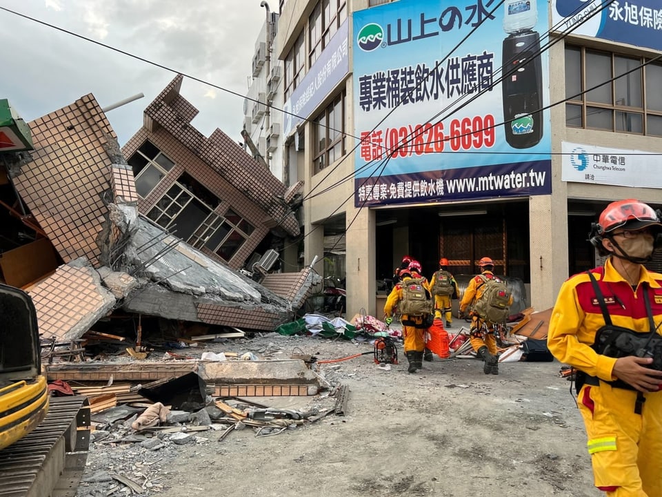 Feuerwehrleute in Schutzanzügen gehen mit Ausrüstung auf ein eingestürztes Gebäude zu.