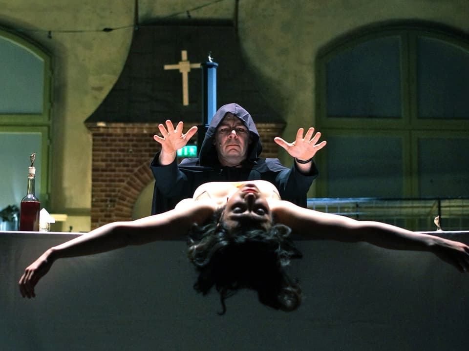 Bühnenszene: Okkultes Ritual mit Priester in schwarzer Kutte und einer Frau, die auf einem Tisch liegt.