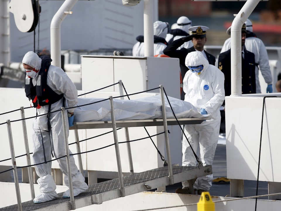 Zwei Männer in weissen Schutzanzügen tragen eine tote Person in einem Weichensack von Bord eines Schiffes.
