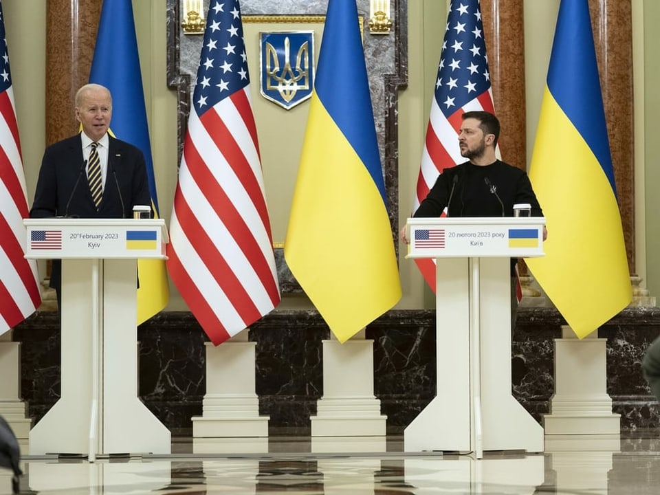 Zwei Männer stehen jeweils vor weissen Rednerpulten. Im Hintergrund sind blau-gelbe sowie rot-weiss-blaue Flaggen zu sehen. 