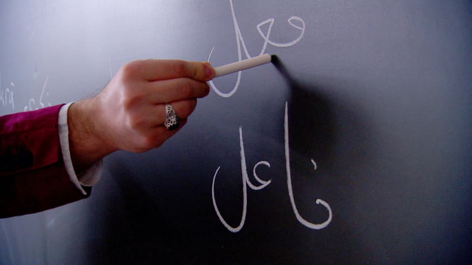 Arabische Schrift auf einer Wandtafel. Eine Hand hält die Kreide.
