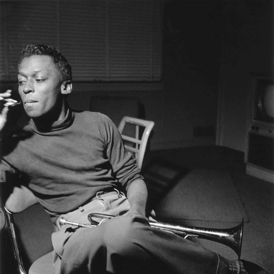 Der junge Miles Davis sitzt auf einem Stuhl. Auf dem Schoss hat er eine Trompete. Er raucht.