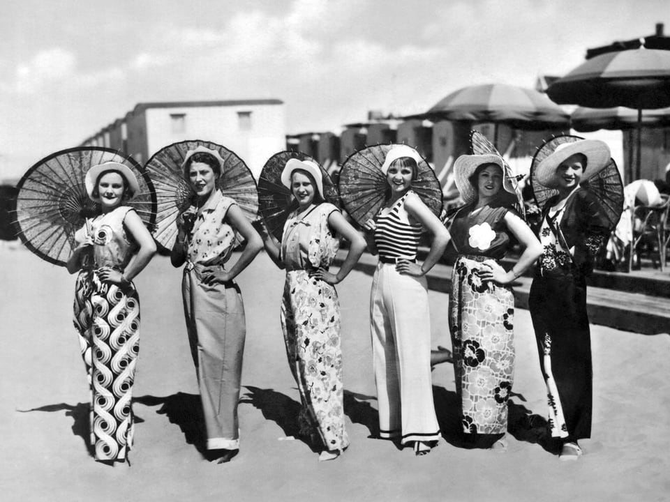 Sechs Frauen in modischen Somemrkleidern mit viel Stoff samt Hüten und Sonnenschirmen