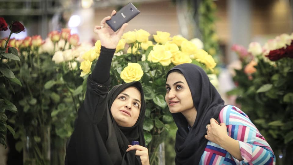 Symbolbild: Zwei junge Iranerinnen mit schwarzem Kopftuch machen vor grossen Blumensträussen mit dem Handy ein Selfie.