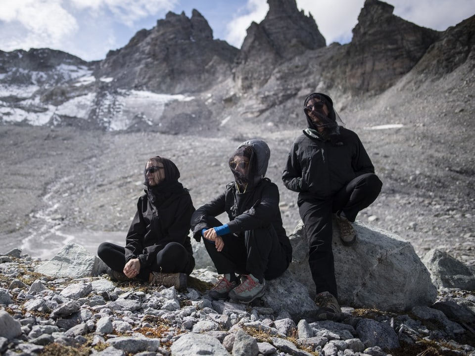 Drei Personen sitzend auf einem Felsen in einer Berglandschaft, bewölkt