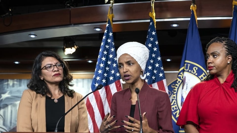 Drei Frauen an Rednerpult. Dahinter US-Fahnen. Eine Frau trägt einen Turban.
