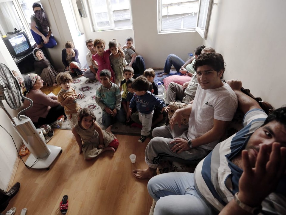 Zahlreiche Kinder, Männer und Frauen sitzen in einem Wohnzimmer