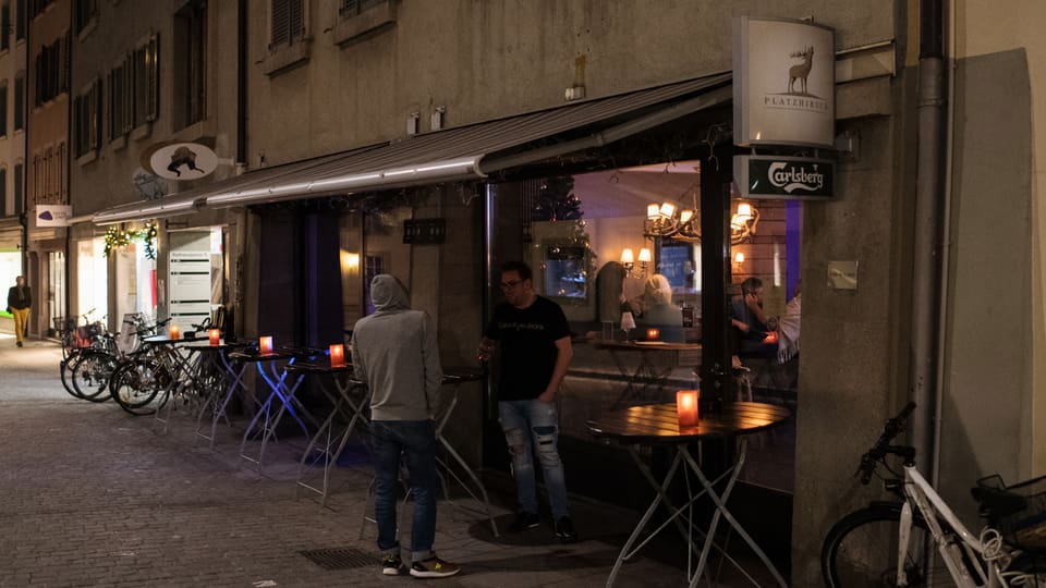 Bar an einer gepflasterten Altstadtgasse. Draussen ist es dunkel, zwei Männer stehen vor der BAr. Durch das Fenster sieht man Menschen im beleuchteten Innenraum.