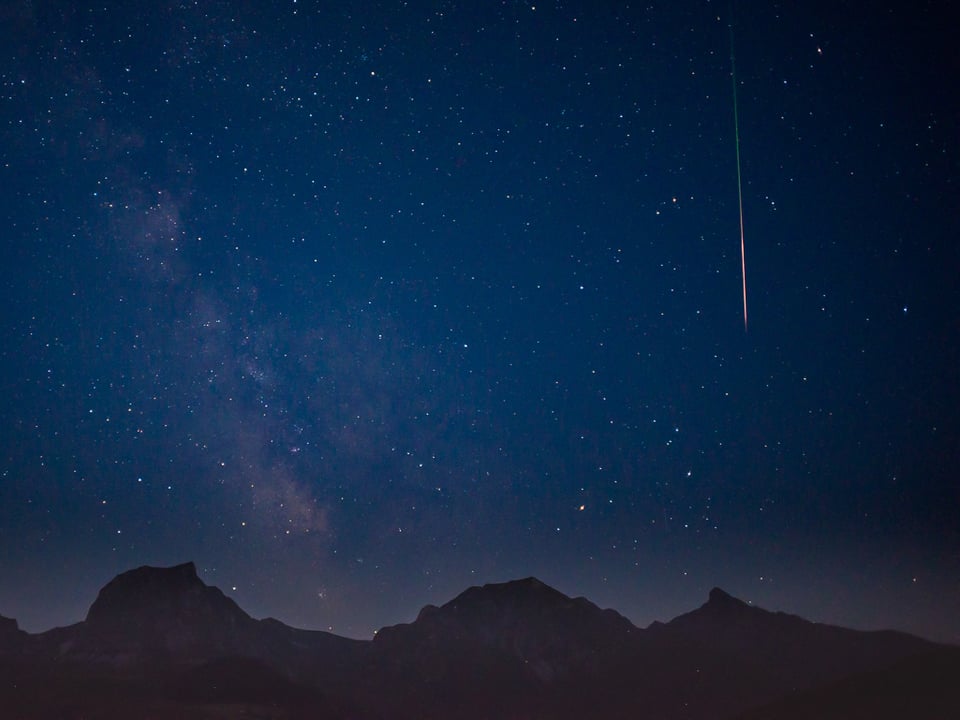 Sternenklarer Nachthimmel mit Sternschnuppe. Unten sind Konturen von Bergen zu sehen. 
