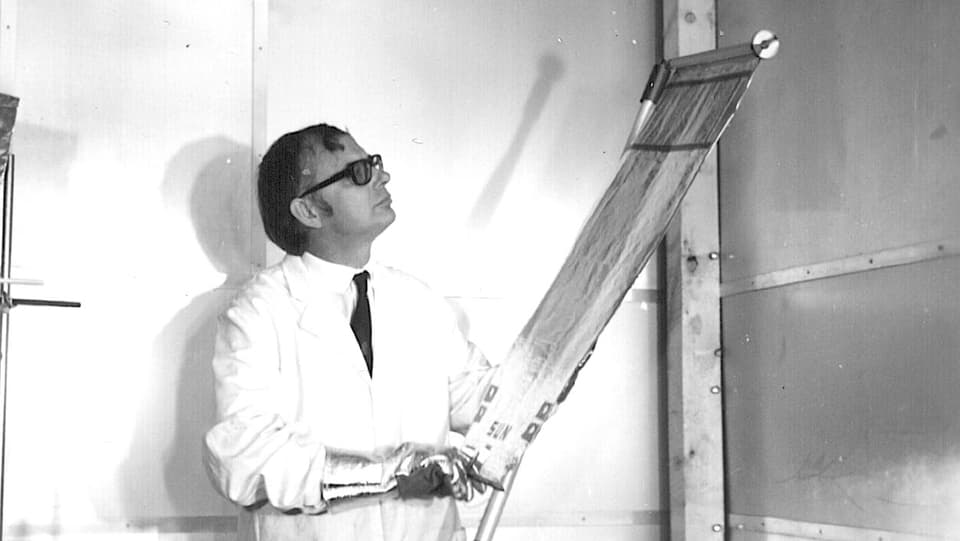 Schwarzweissfoto: Mann in weissem Laborkittel zieht ein silbern glänzendes Rollo auf.
