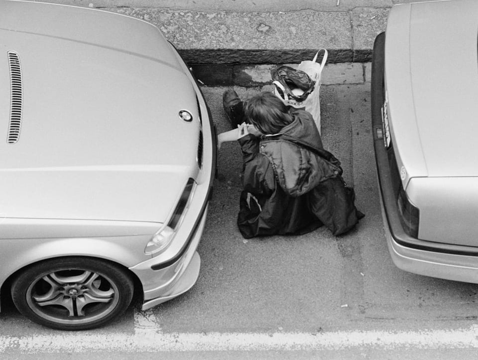 Schwarzweiss-Foto: Drogensüchtige Person zwischen zwei parkierten Autos.