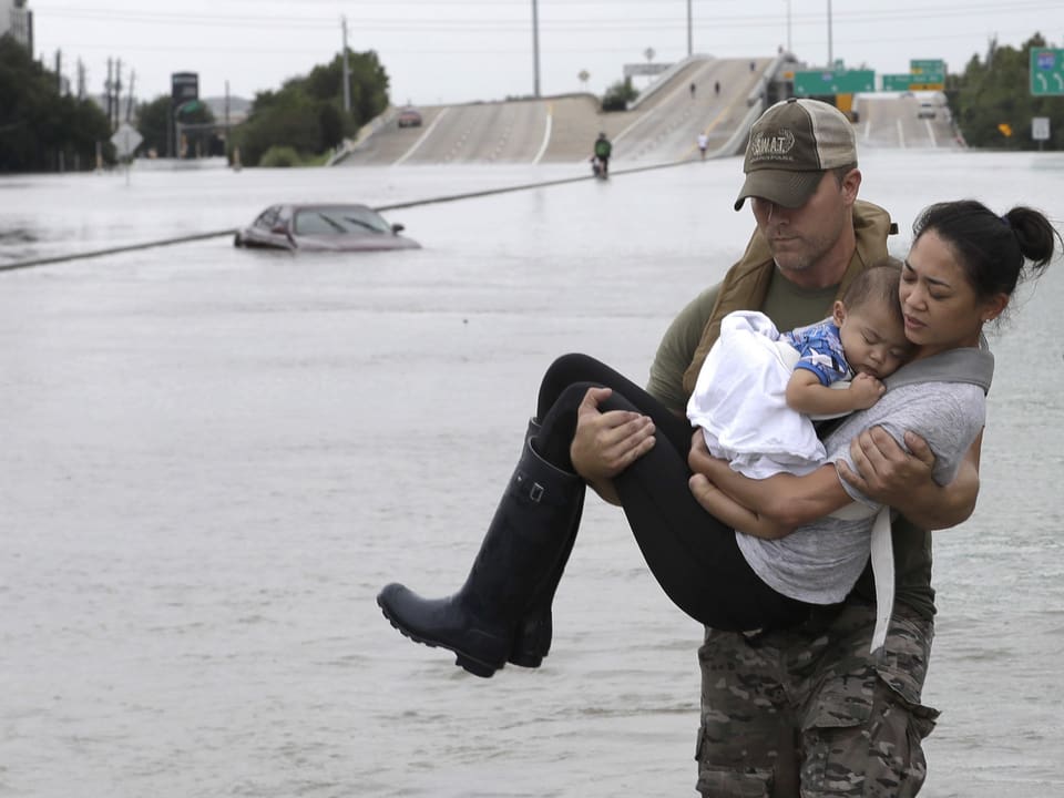 Ein Mann trägt eine Frau durch das Wasser. Diese hat ihren Säugling im Arm.