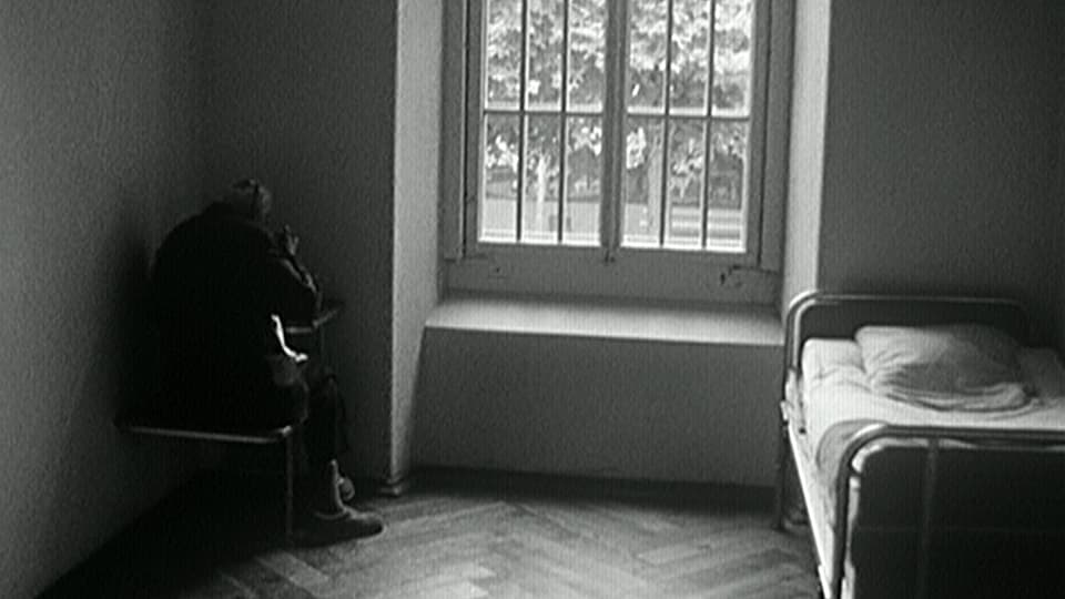 Schwarz-weiss-Bild eines Klink-Zimmers. Eine Frau sitzt in der Ecke an einem Tischchen.