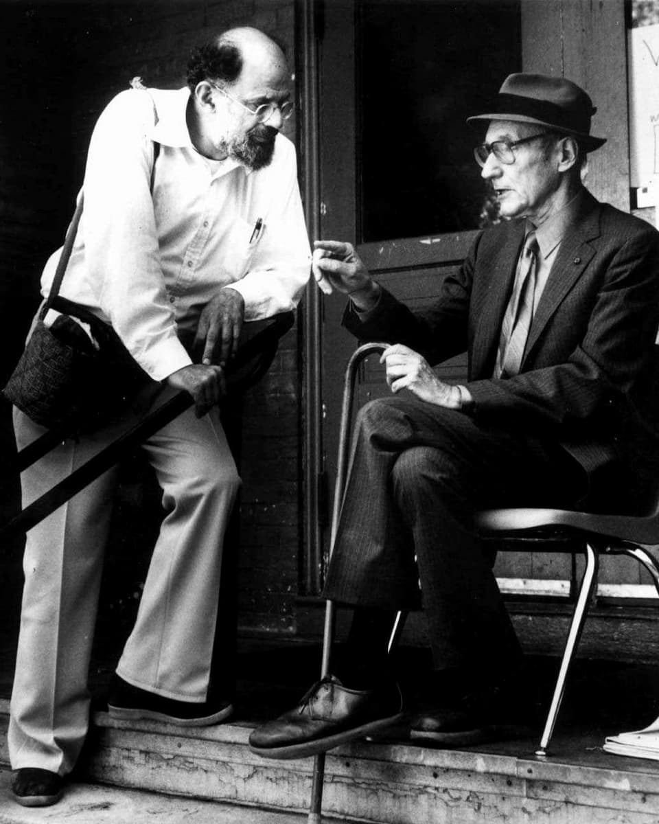 Allen Ginsberg steht und redet mit Burroughs, der auf einem Stuhl sitzt. 