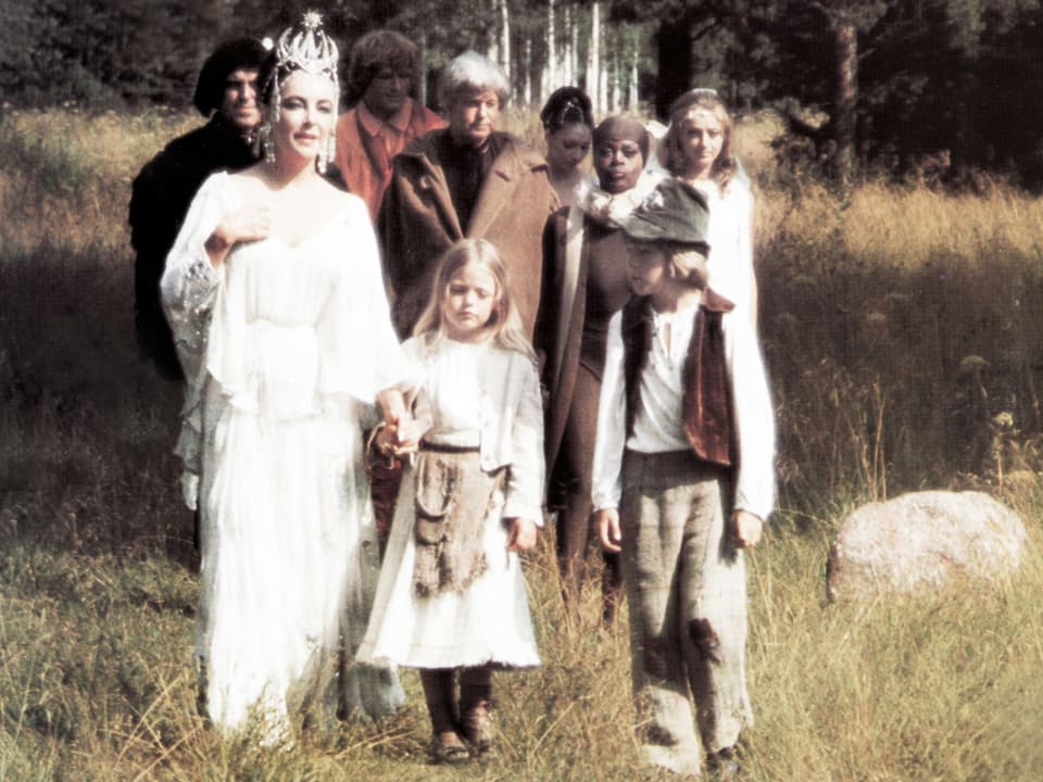 Eine Frau in Weiss hält ein kleines Mädchen bei der Hand, daneben laufen andere Menschen durch die Natur.
