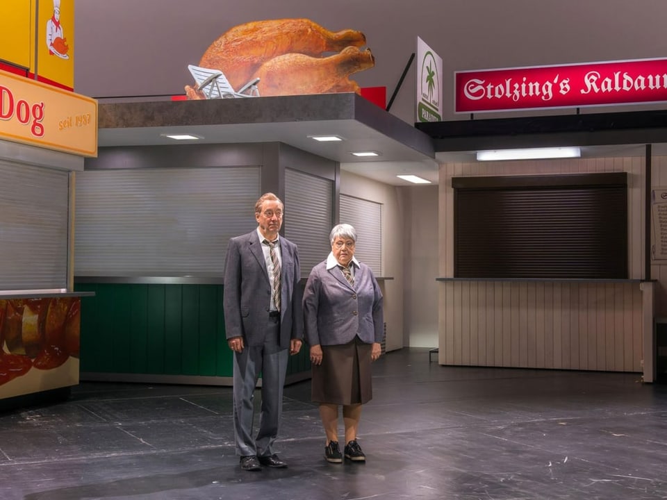 Ein Passant und eine Passantin auf der Theaterbühne zwischen Würstchenbuden.