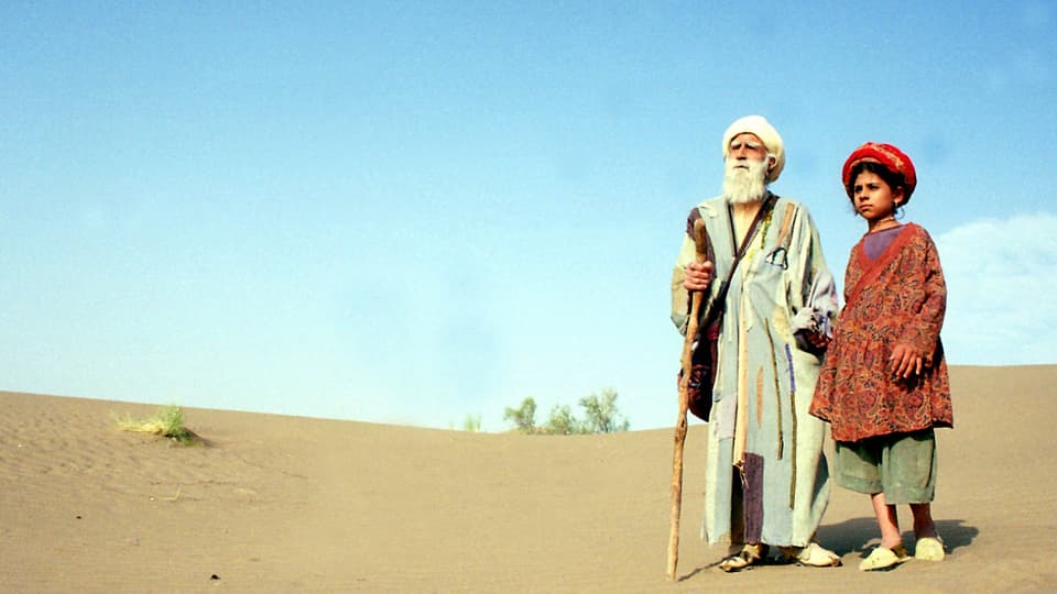 An old man and a young woman, both in oriental dresses, stand in the desert