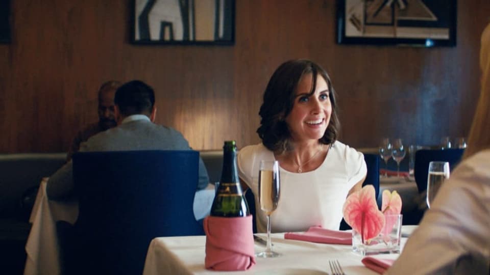 Filmszene: Eine Frau sitzt in einem Restaurant am Tisch und lacht, vor ihr eine Flasche Champagner.