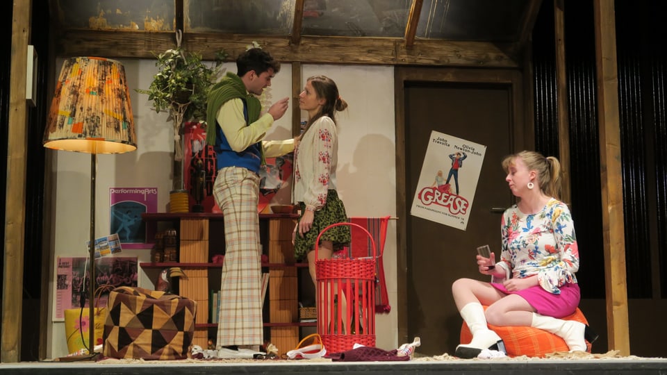 Szene in einem Landtheater: Ein Paar diskutiert stehend auf der Bühne, daneben sitzt eine junge Frau auf einem Hocker.