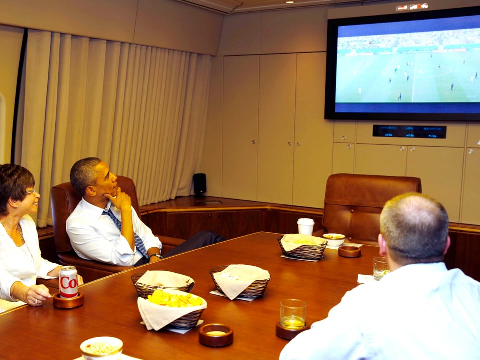 Barack Obama schaut mit zwei Personen in der «Air Force One» ein Fussballspiel im Fernsehen.
