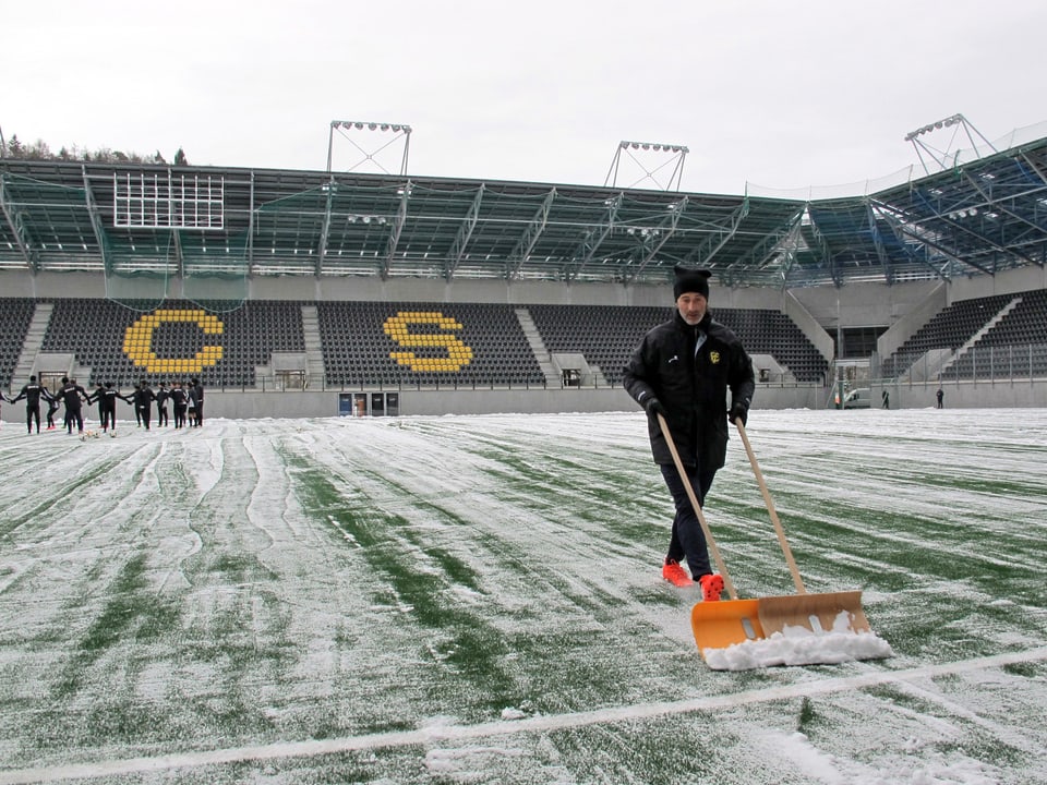 Ein Mann mit organgen Schuhen und schwarzem Trainingsanzug schiebt Schnee mit einer grossen Schaufel von einem Fussballfeld.