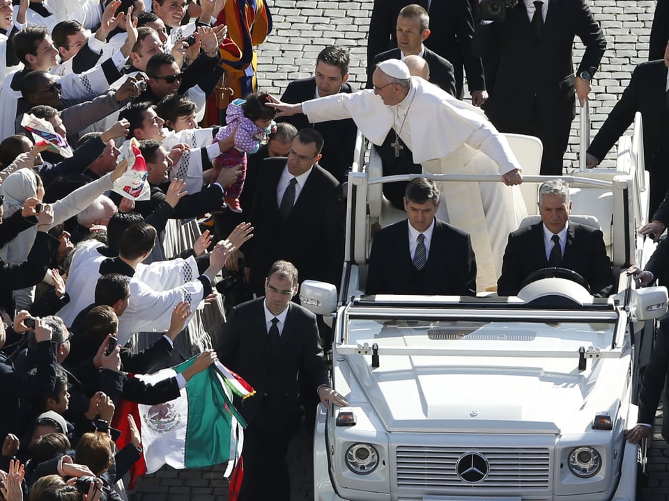 Papst Franziskus im offenen Wagen, ein Kind segnend.
