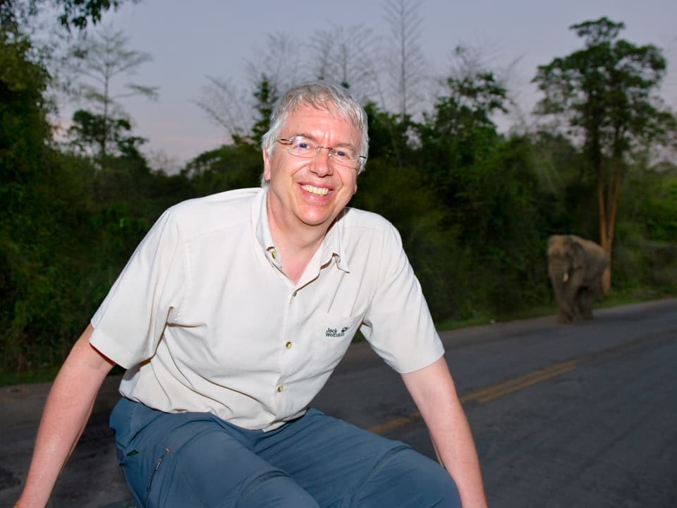 Zoo-Direktor Alex Rübel posiert im Abendlicht, im Hintegrund spaziert ein Elefant der Landstrasse entlang.
