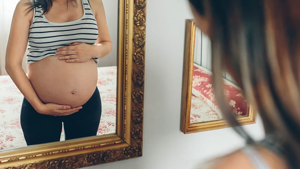 Eine schwangere Frau betrachtet sich im Spiegel.