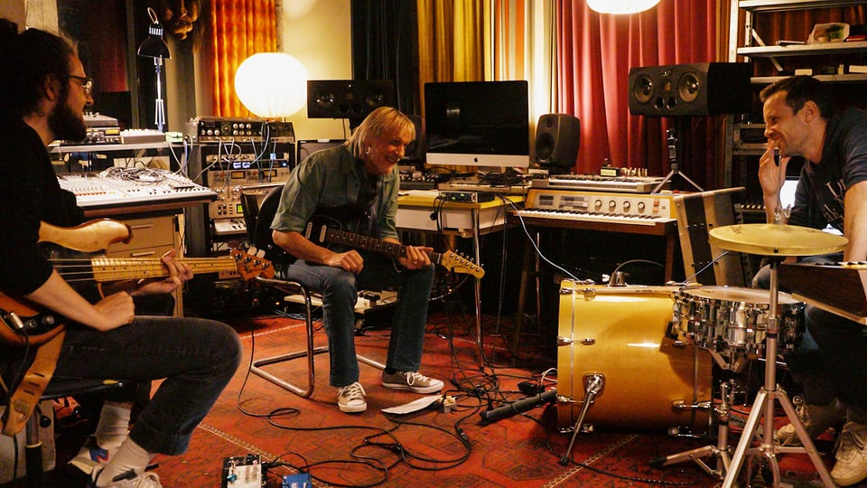 Man playing guitar in a music study. At the edge of the picture, younger men with bass and drums.