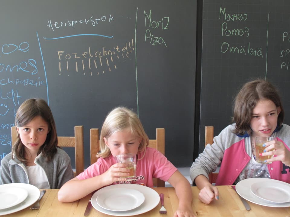 Drei Mädchen sitzen vor leeren Tellern am Tisch, hinten auf der Wandtafel steht Fozelschnitten!!!!! oder Piza