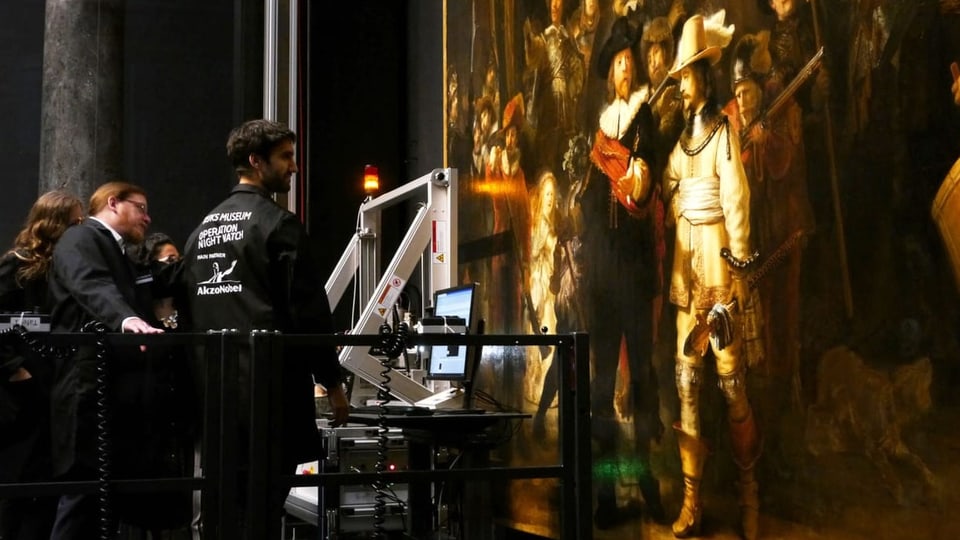 Menschen hantieren an technischen Geräten, die vor einem grossen Gemälde mit zahlreichen Personen drauf stehen.