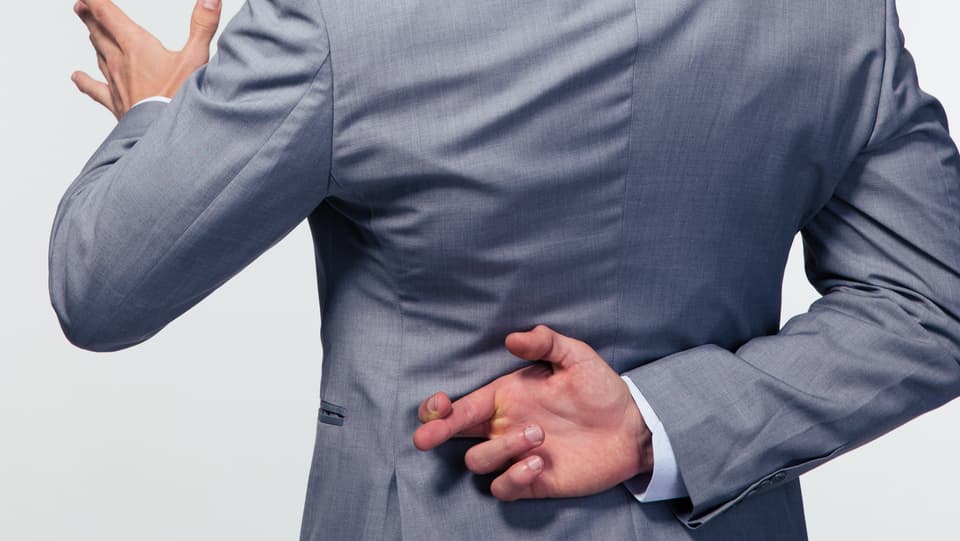 Ein Mann in Anzug spricht und überkreuzt hinter seinem Rücken die Finger. 
