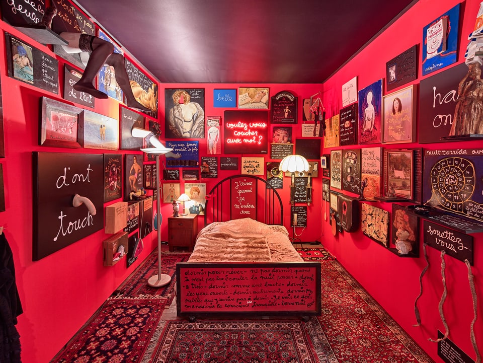 Ein rot gestrichener Raum mit einem Bett in der Mitte. An den Wänden hängen erotische Werke und Schriftbilder. 