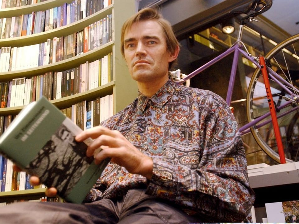 Zimmermann trägt ein farbiges Hemd, und hält sein Buch in den Händen. Er sitzt in einem Zürcher Buchladen, im Schaufenster steht ein violettes Rennvelo.