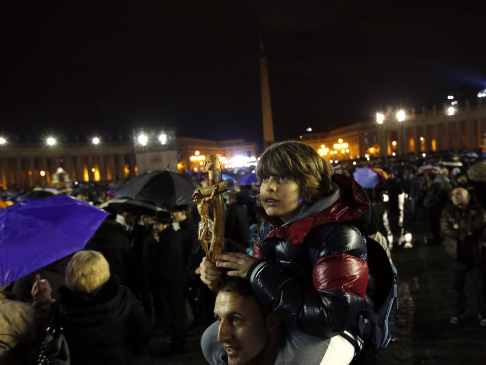Junge auf den Schultern seines Vaters mit Kruzifix in der Hand auf dem Petersplatz. Nacht.