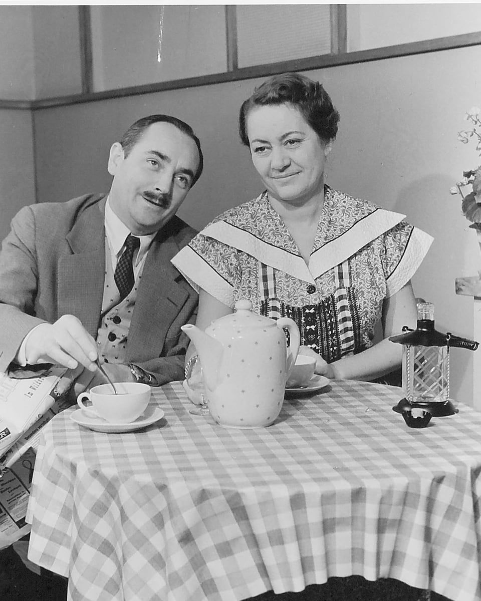 Schwarz-weiss Fotografie. Die Schauspieler sitzen am Tisch, auf dem zwei Tassen und eine Kaffeekanne steht.