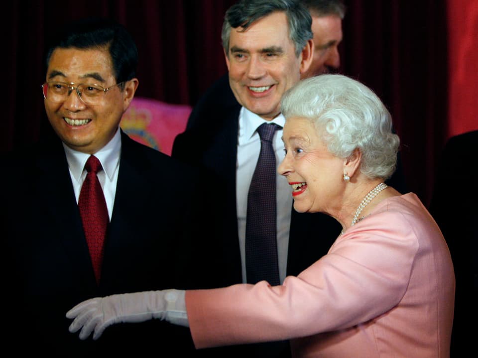 Die Queen im lachsfarbenen Kostüm streckt ihre Hand jemandem entgegen, der nicht auf dem Foto sichtbar ist. Hinter ihr stehen zwei lachende Männer in Anzügen.