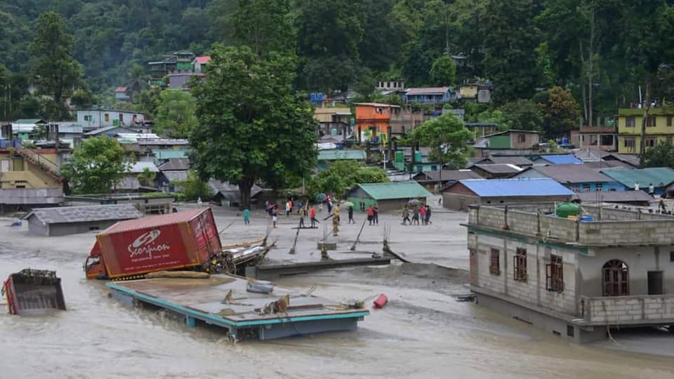 Menschen stehen auf der Strasse eines überfluteten Dorfes, ein LKW wurde von den Wassermassen weggeschwemmt.