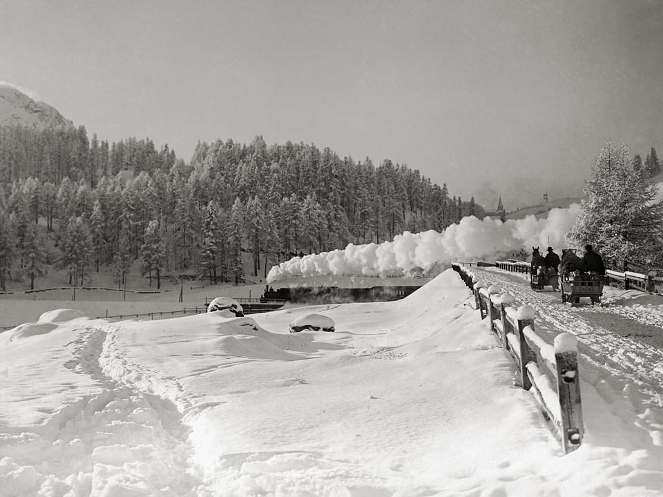 Das Foto zeigt einen historischen Zug unter Dampf im Hintergrund, während im vorderen Teil zwei Pferdekutschen unterwegs sind.