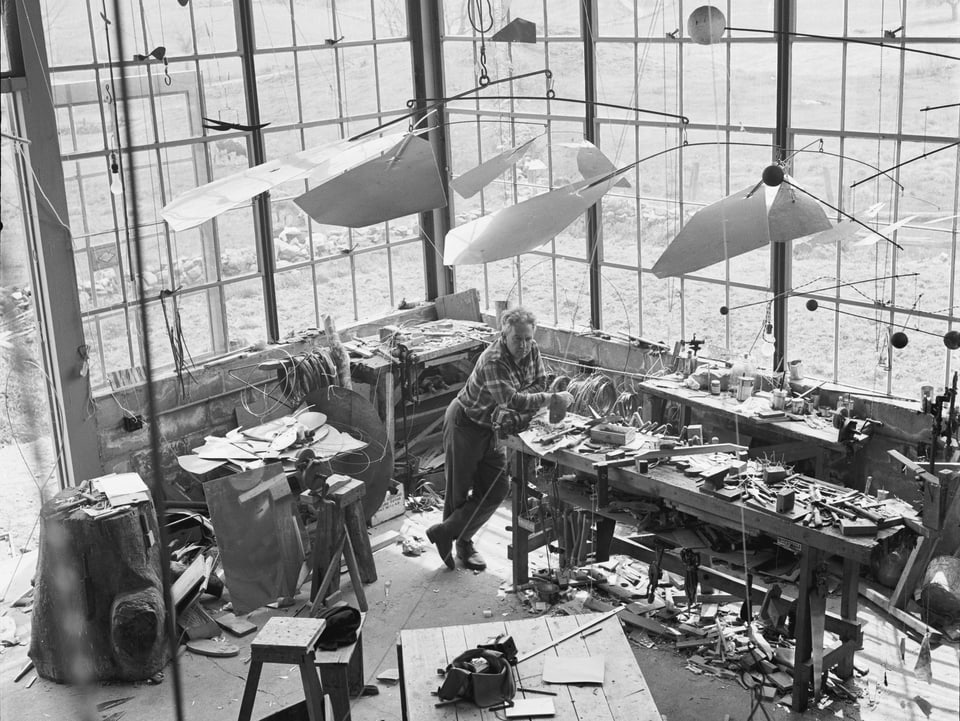 Calder steht in seinem Atelier. Bild aus der Vogelperspektive. Im Atelier hängen viele Mobilés.
