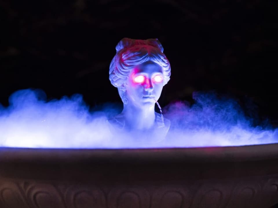 Büste einer Statue mit leuchtenden roten Augen und bläulicher Beleuchtung in Nebel.