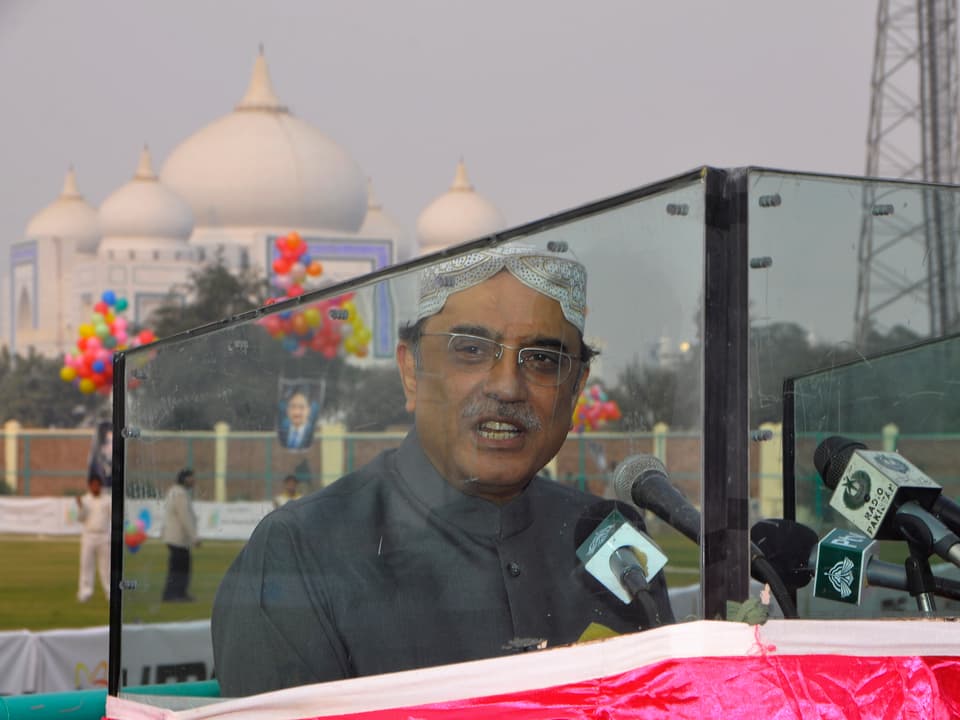 Zardari bei einer Rede hinter Panzerglas, im Hintergrund ein Mausoleum.