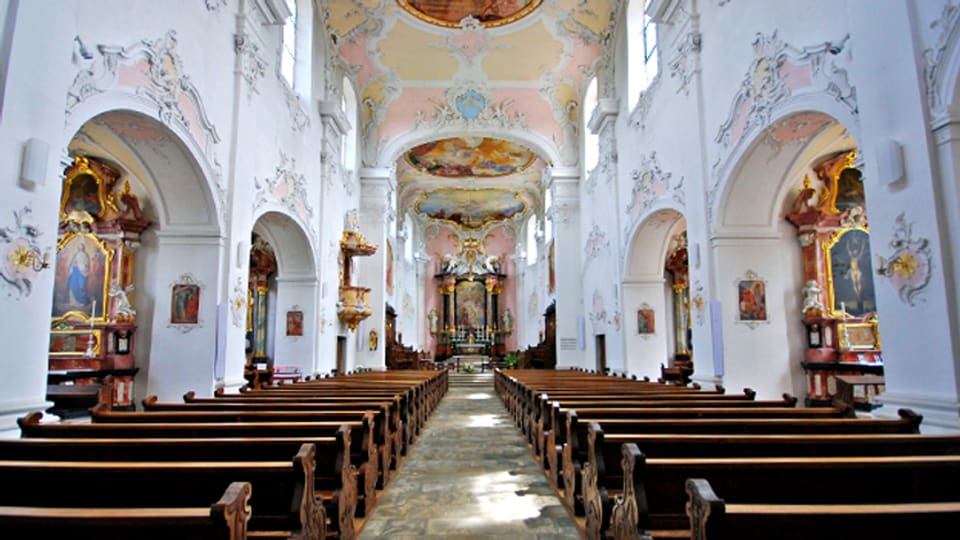 Der Innenraum einer barocken Kirche: dunkle Holzbänke, bunte Deckenfresken, gold-verzierte Altare.