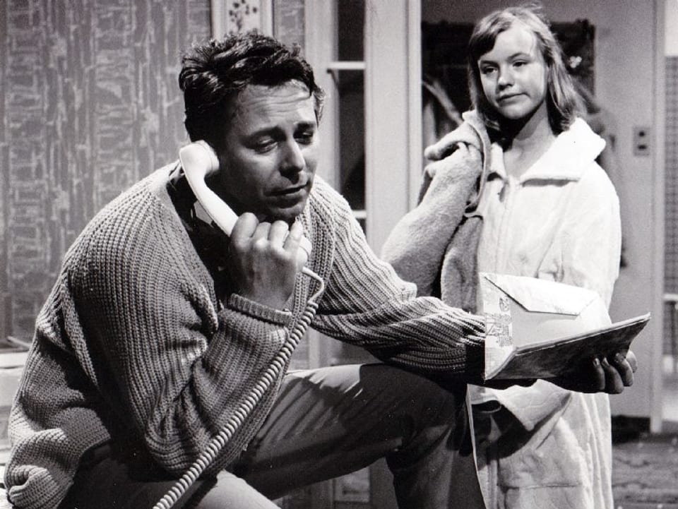 Ein Mann (sitzend) telefoniert mit der rechten Hand. In der Linken hält er zwei Briefumschläge. Hinter ihm steht ein Mädchen im Bademantel. 