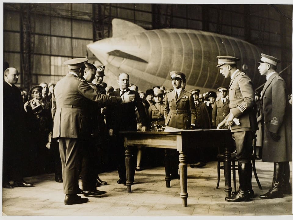Vergilbte Schwarzweiss-Aufnahme: Männer in Uniform stehen um Tisch, im Hintergrund ein Zeppelin